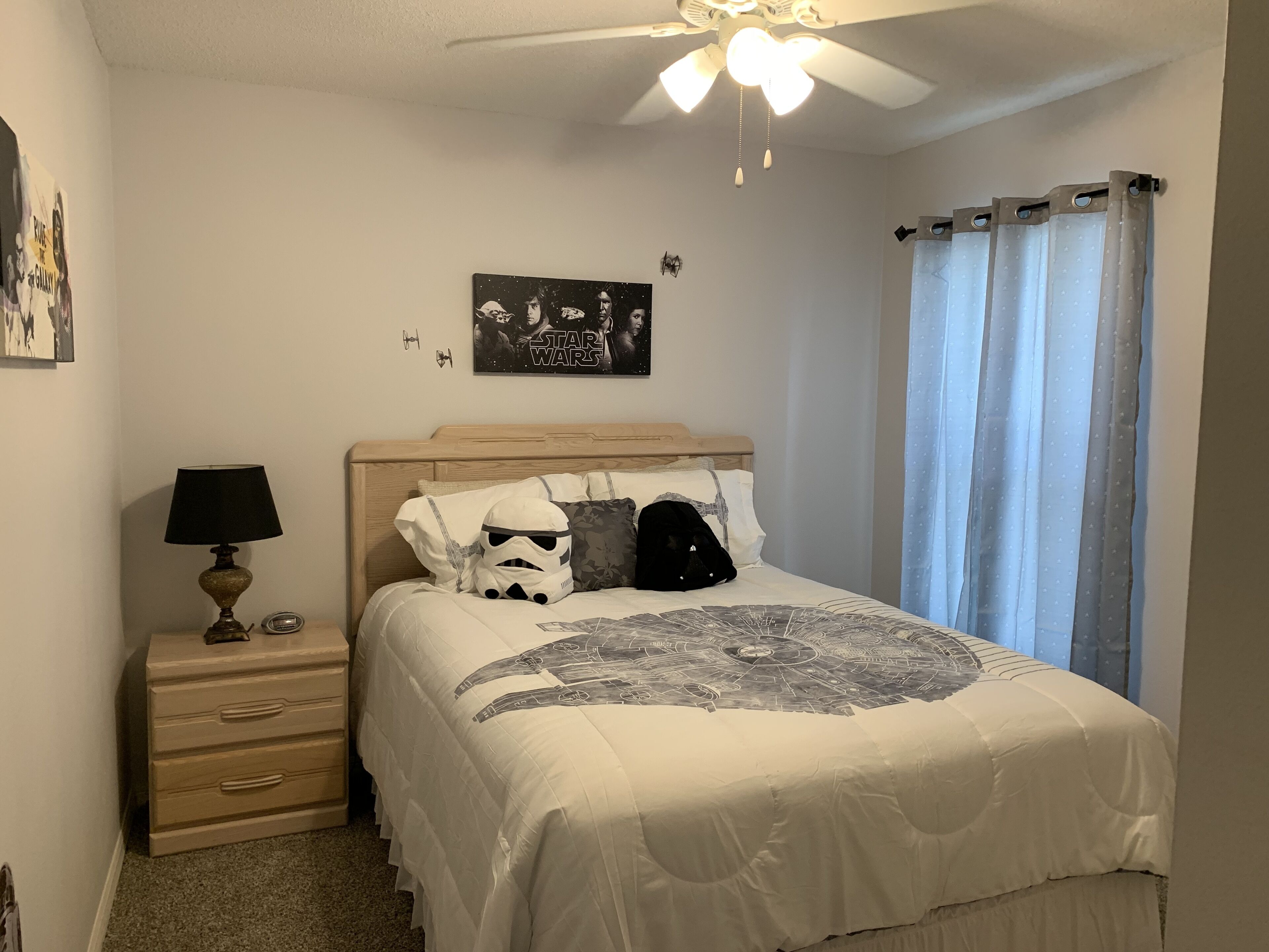 A bedroom with a bed, a nightstand, a lamp, a Star Wars poster, and a ceiling fan.