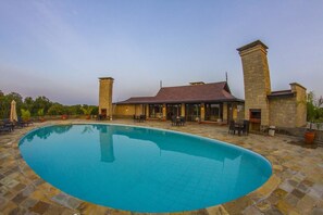 Une piscine avec une terrasse en pierre, un espace extérieur couvert aménagé en salon, et un bâtiment doté d’un toit rouge.