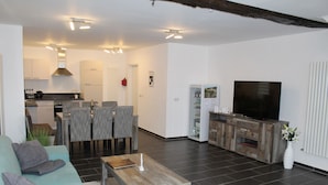 Ein modernes Wohnzimmer mit Sofa, Couchtisch, Fernseher und einem Essbereich mit Stühlen sowie einer Küche im Hintergrund.