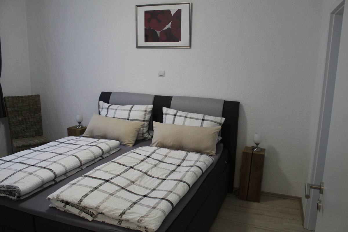 Una cama doble con una colcha y almohadas a cuadros, una mesita de noche con lámpara y un cuadro en la pared.