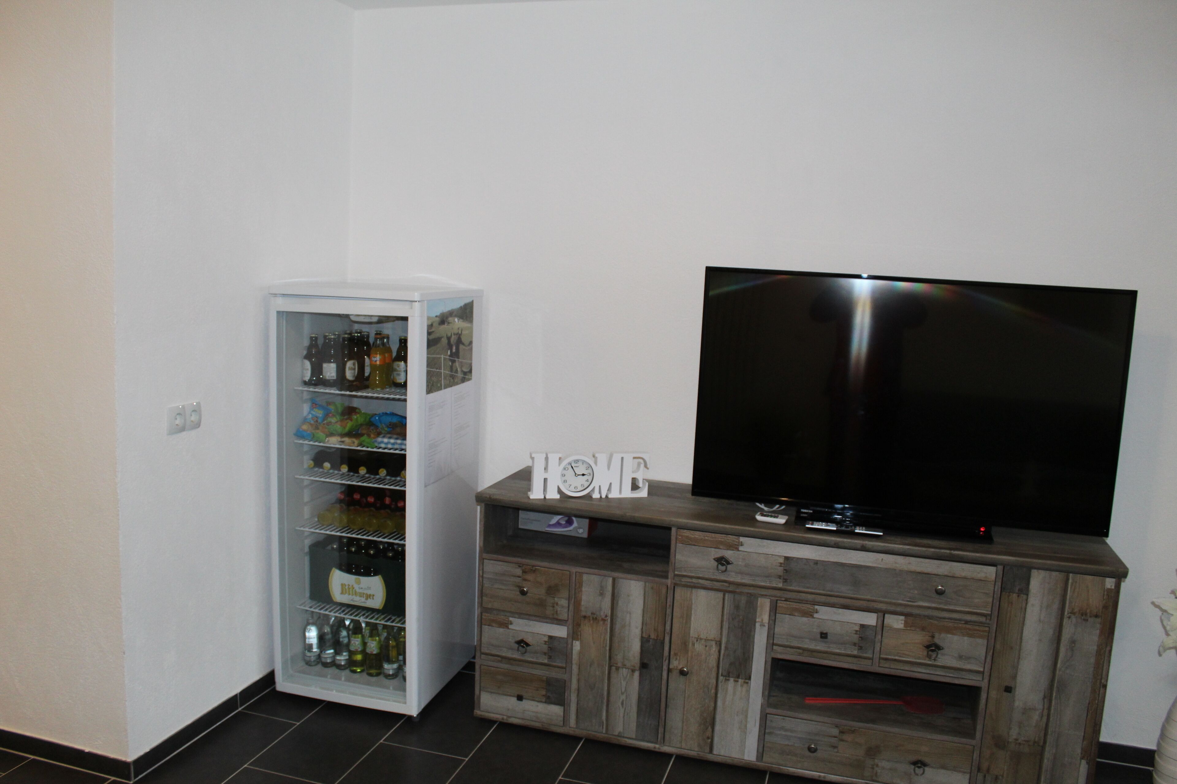 A living room with a flat-screen TV on a wooden cabinet, a mini-fridge with beverages, and a 'HOME' sign on the cabinet.