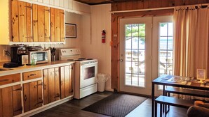 A cozy cabin-style kitchen with wooden cabinets, a stove, and a microwave. There is a dining table with a bench and a view of the outdoors through a glass door. - Amenities & Resort Access Included, Cabin 11 (Phelps)
