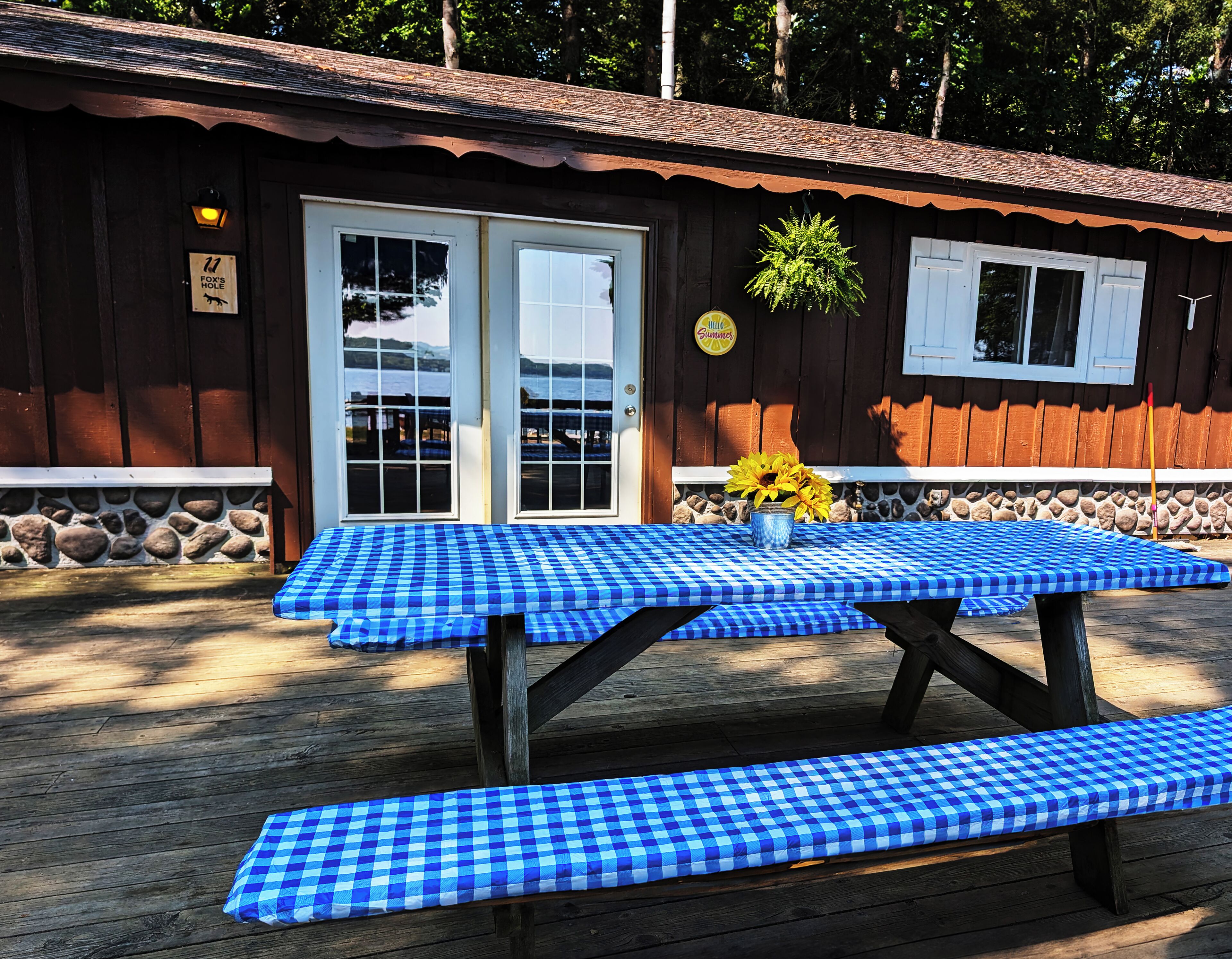 A wooden cabin with a checkered picnic table and bench, a stone wall, and a sign reading "Fox's Hole".
