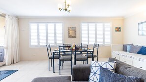 Un salon moderne avec un coin repas, comprenant un lustre, de grandes fenêtres équipées de stores, un canapé agrémenté de coussins à motifs et une petite table avec une bouteille et des verres.