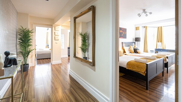 A hotel room with a wooden floor, a large mirror, a bed with a yellow blanket, and a potted plant. - Nice 3 Bedrooms Apartment Near Hyde Park & Oxford St (London)