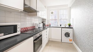 A modern kitchen with white cabinets, a microwave, a washing machine, and a window with frosted glass. - Nice 3 Bedrooms Apartment Near Hyde Park & Oxford St (London)
