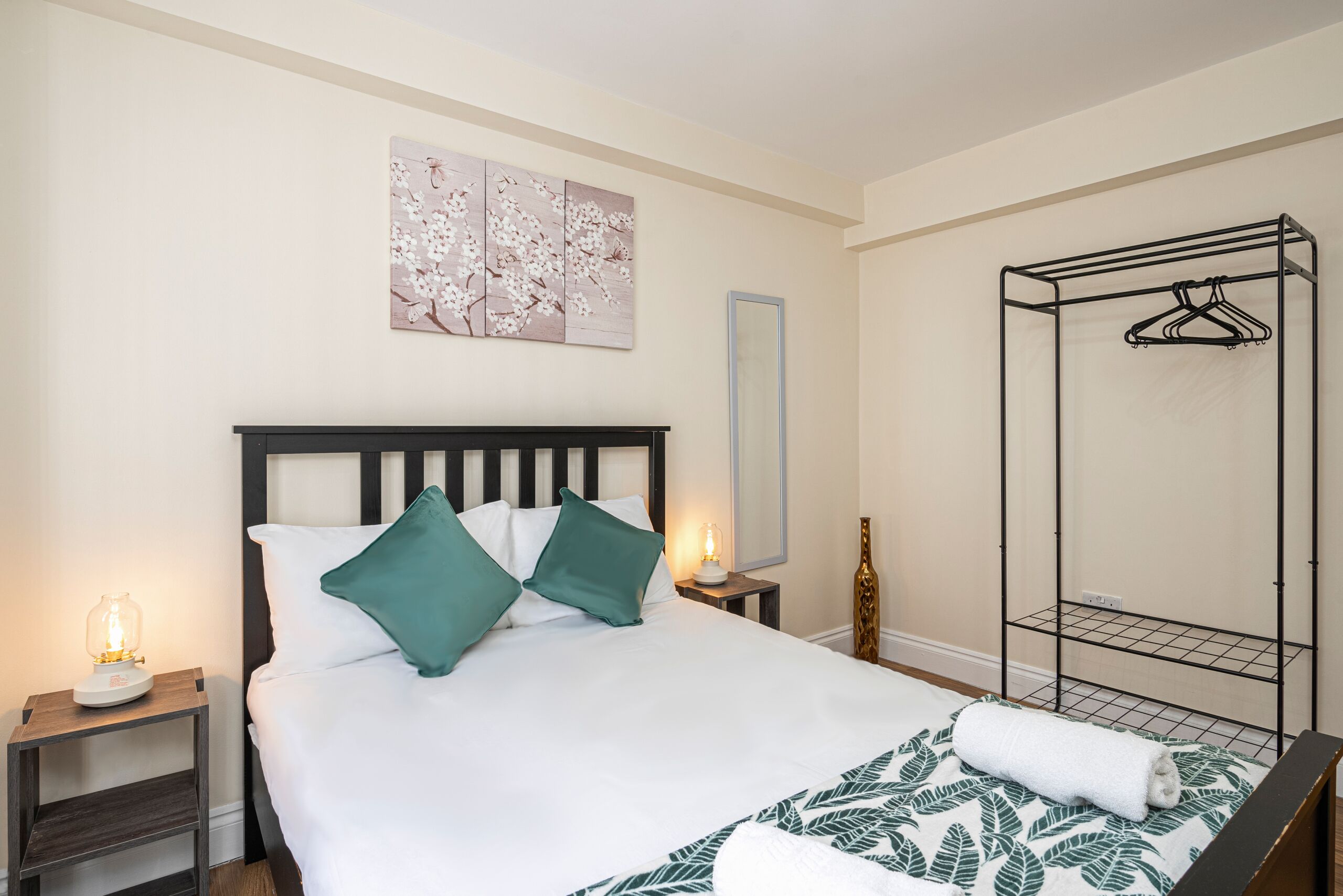 Een netjes opgemaakt bed met wit en groen beddengoed, een nachtkastje met een lamp, een wandspiegel en een metalen rek.