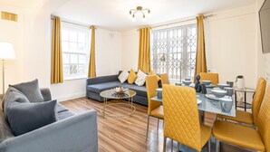 A modern living room with a sofa, a dining table with chairs, and large windows with curtains. - Nice 3 Bedrooms Apartment Near Hyde Park & Oxford St (London)