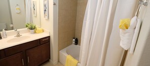 Ein zweites Badezimmer mit Badewanne, Duschvorhang, Waschbecken mit Pflegeartikeln und Spiegel.