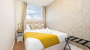 A hotel room with a bed, a nightstand, a lamp, and a window with curtains. - Lovely Apartments in Landward Court (London)