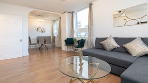 A modern living room with a glass coffee table, grey sofa, and a dining area with chairs. - Lovely Apartments in Landward Court (London)