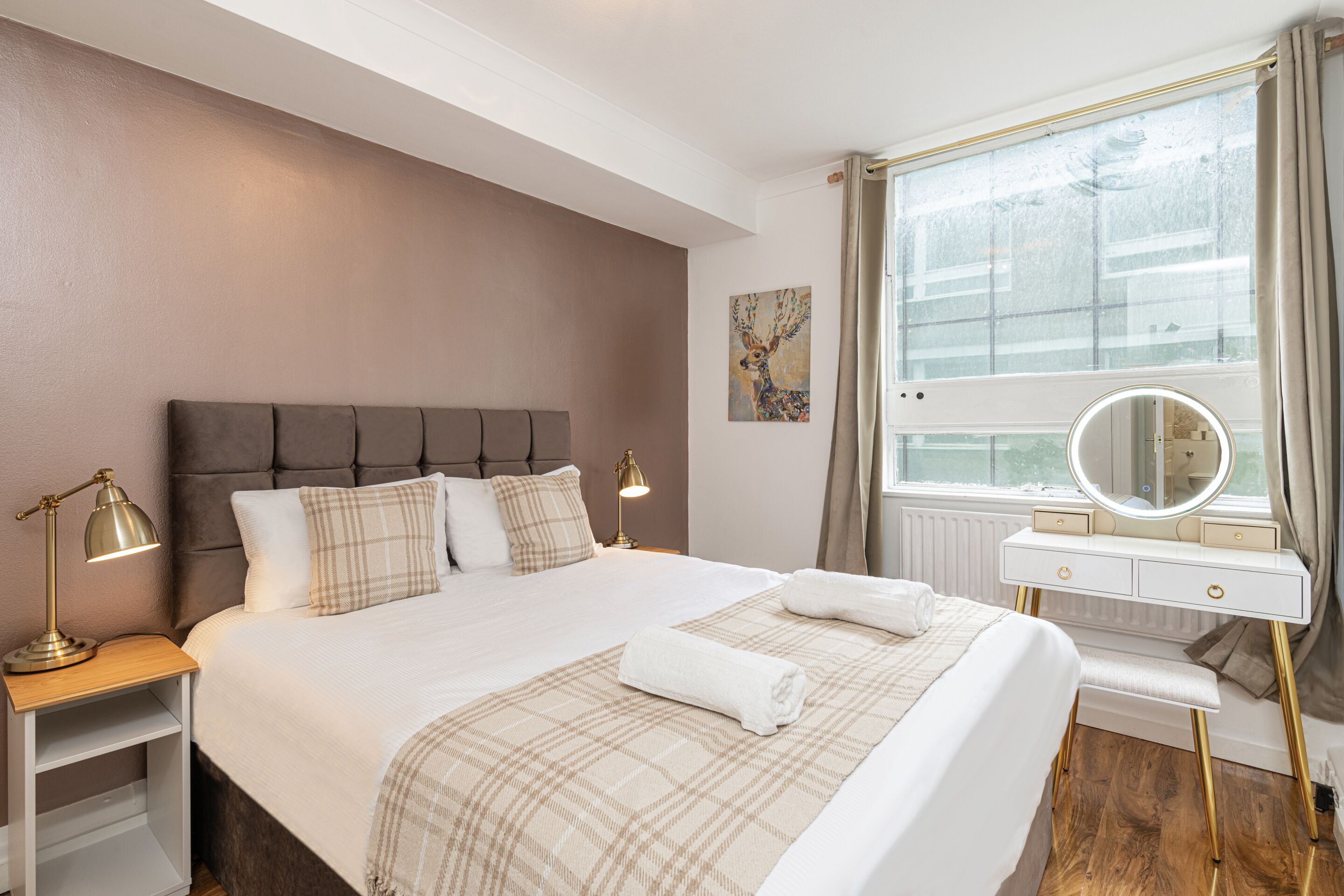 A neatly arranged hotel room with a large bed, bedside lamps, a vanity with a mirror, and a window with curtains.