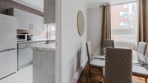 A modern kitchen with white cabinets, a microwave, and a coffee maker. There is a dining area with a glass table and cushioned chairs. - Lovely Apartments in Landward Court (London)