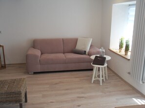 Uma sala de estar com sofá, uma mesa redonda pequena com um livro, uma cadeira de vime e uma janela com plantas.
