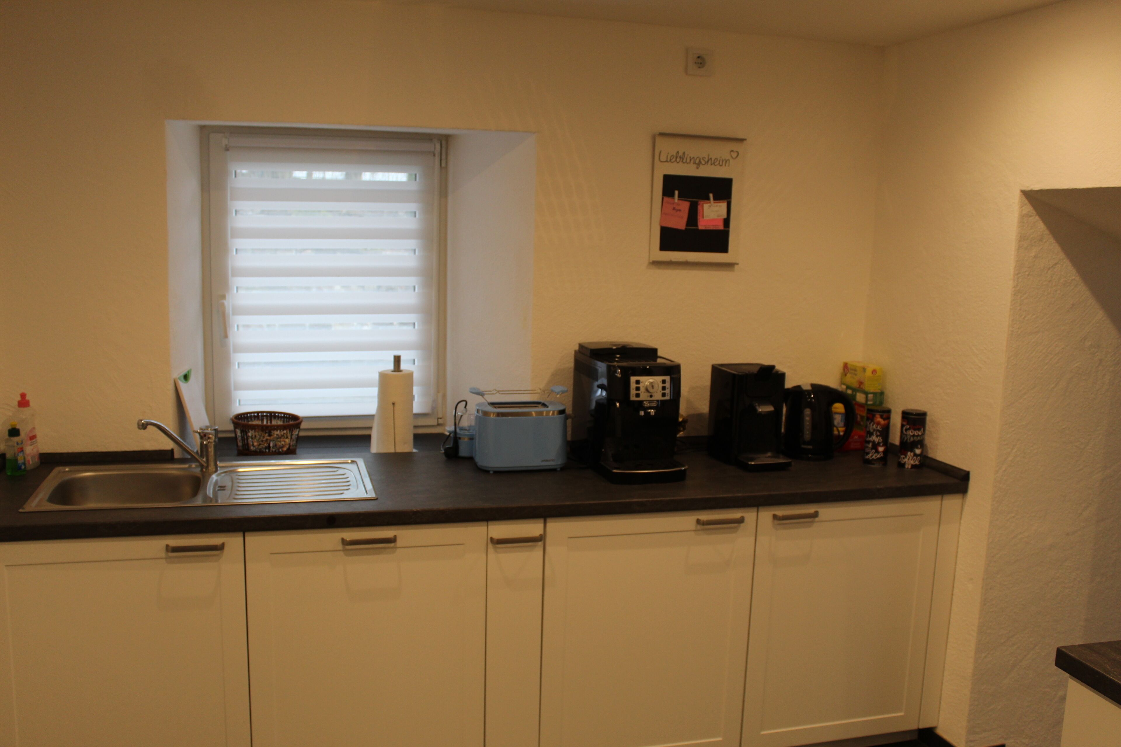 Eine kleine Küche mit Spüle, Toaster, Kaffeemaschine und einem Fenster mit Jalousien.