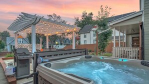 Ein Whirlpool unter einem überdachten Pergola, umgeben von einer Holzterrasse und einem Haus.