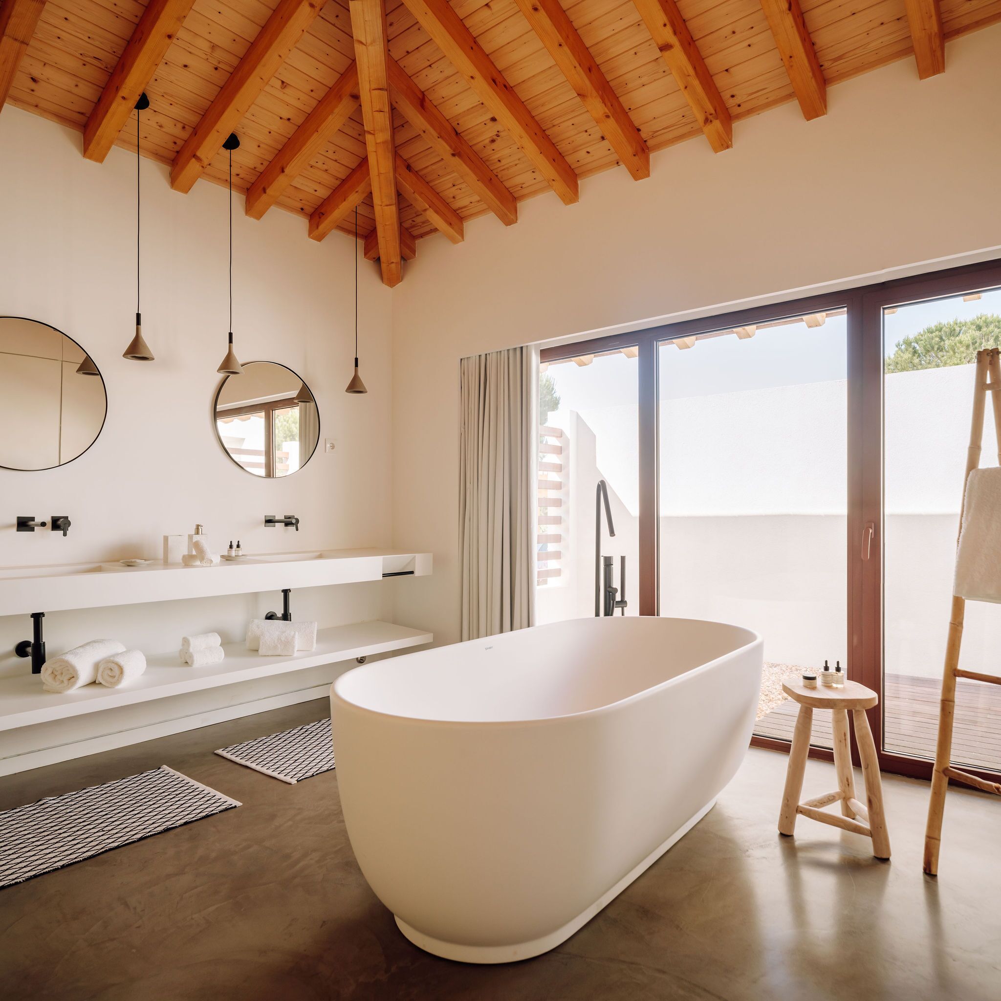 A modern bathroom with a freestanding bathtub, wooden ceiling beams, and a large window offering a view of the outdoors.