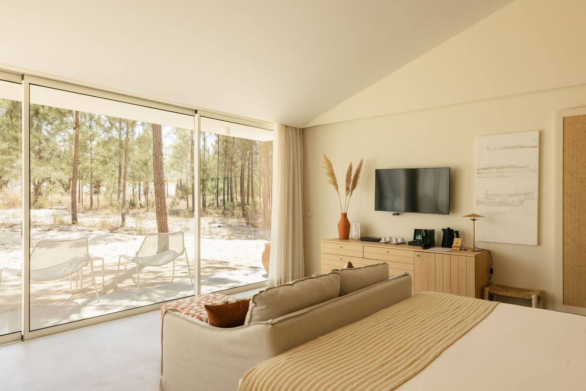 A modern bedroom with a large sliding glass door leading to an outdoor area, a bed with a striped blanket, a TV mounted on the wall, and a wooden dresser with a vase of pampas grass.