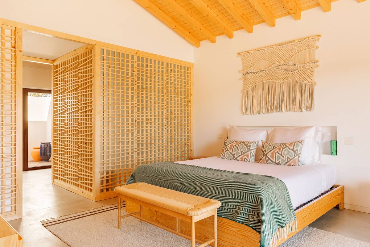 A bedroom with a wooden bed frame, a bench, a woven partition, and a macramé wall hanging.