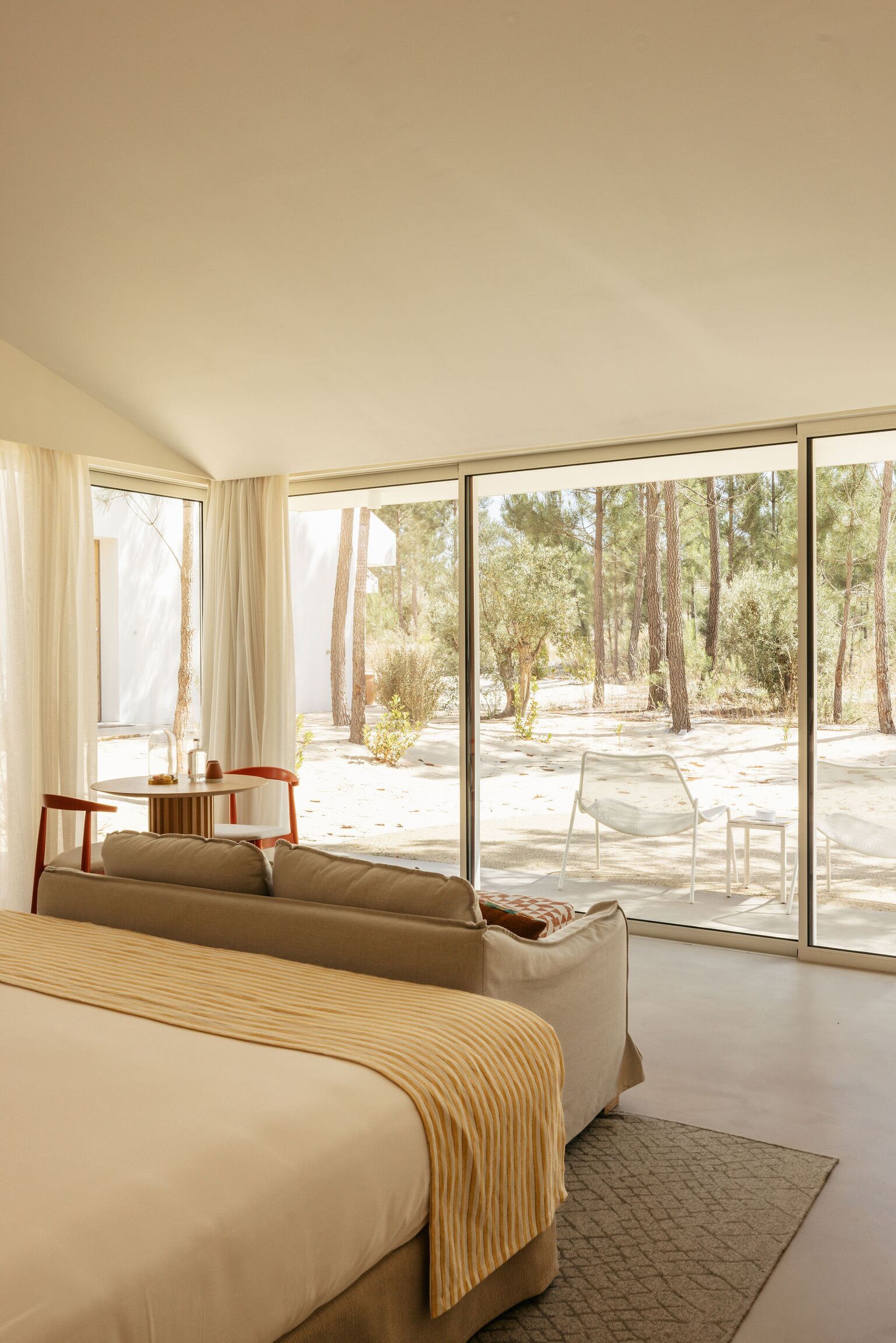 A bedroom with a large bed, a seating area with a table, and a view of a forest through large windows.