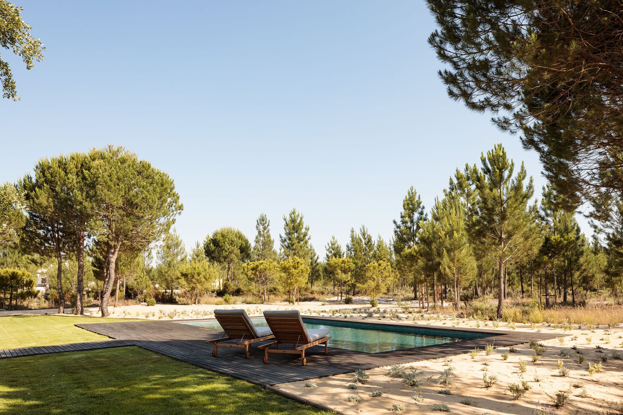 A pool area with two lounge chairs, surrounded by a wooden deck and a sandy landscape with trees.