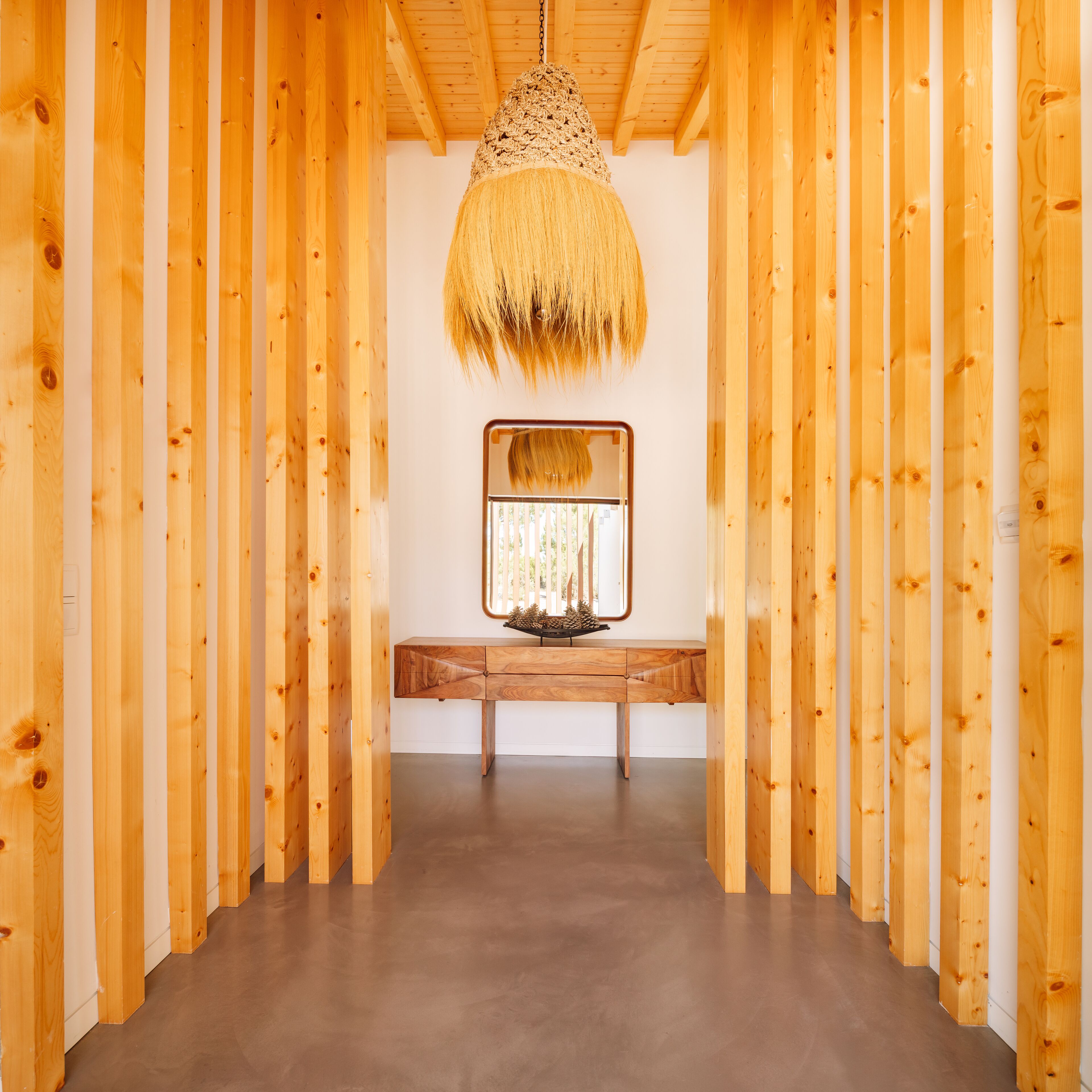 A hallway with wooden walls and a ceiling, leading to a bench with a mirror and a small table with pinecones.
