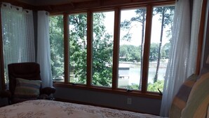 Ein Schlafzimmer mit großem Fenster, einem Bett mit Blumenmuster-Überwurf, einem Sessel mit gestreiftem Kissen und Blick auf einen See und Bäume.