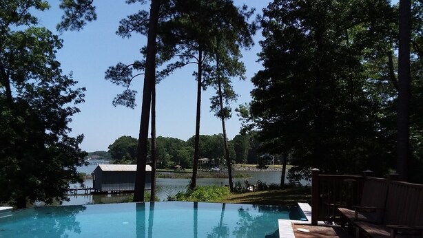 Ein Schwimmbecken im Freien mit einer Holzterrasse und Bänken, umgeben von hohen Bäumen und mit Blick auf einen See sowie Gebäude im Hintergrund.