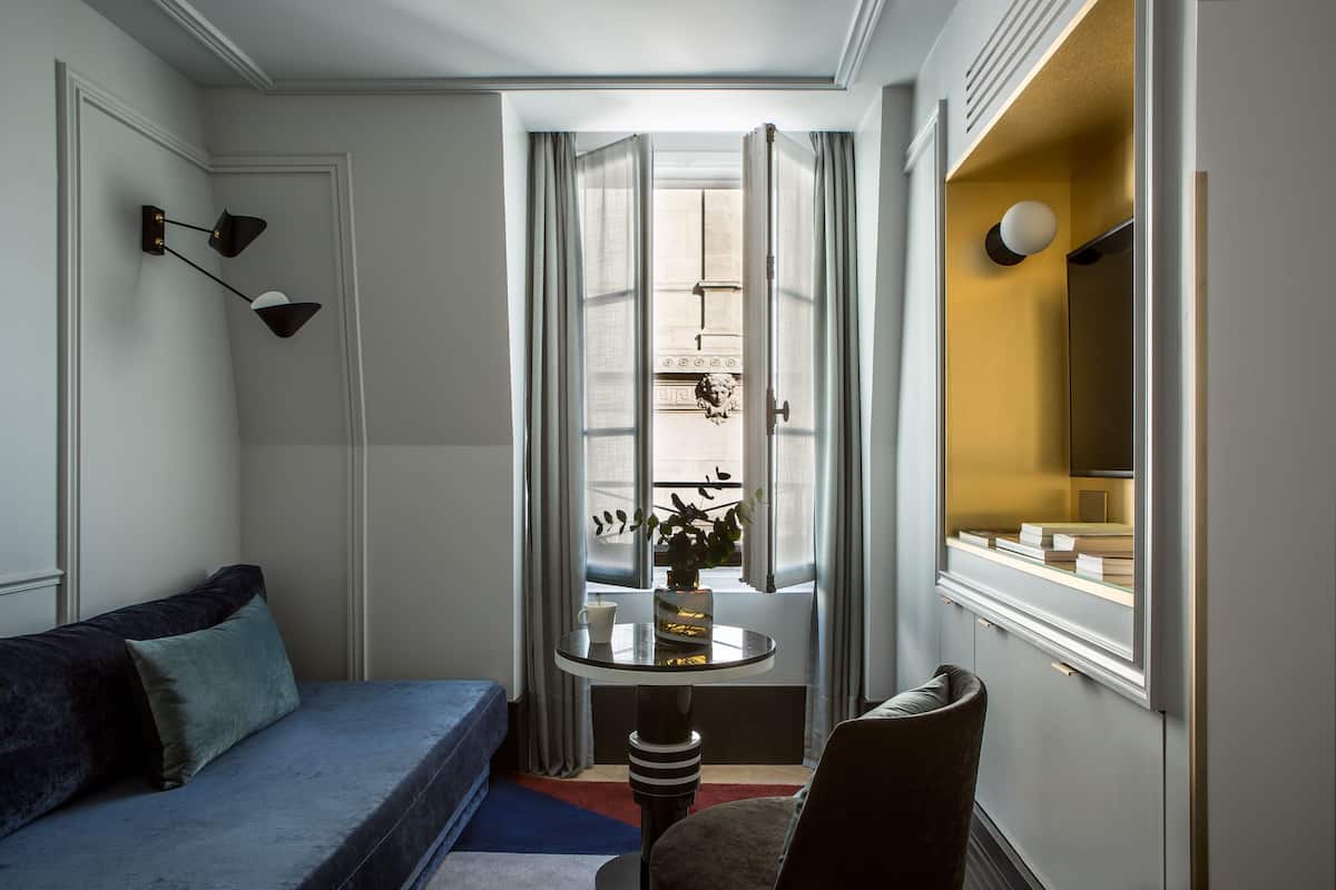 A hotel room with a blue sofa, a small round table with a vase, a chair, a mirror, and a view of the street outside.