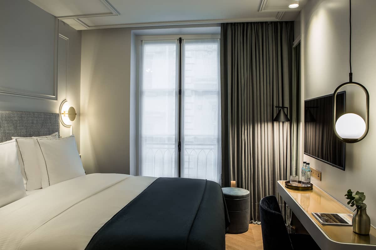 A hotel room with a bed, bedside table, mirror, and a view of the city through large windows.