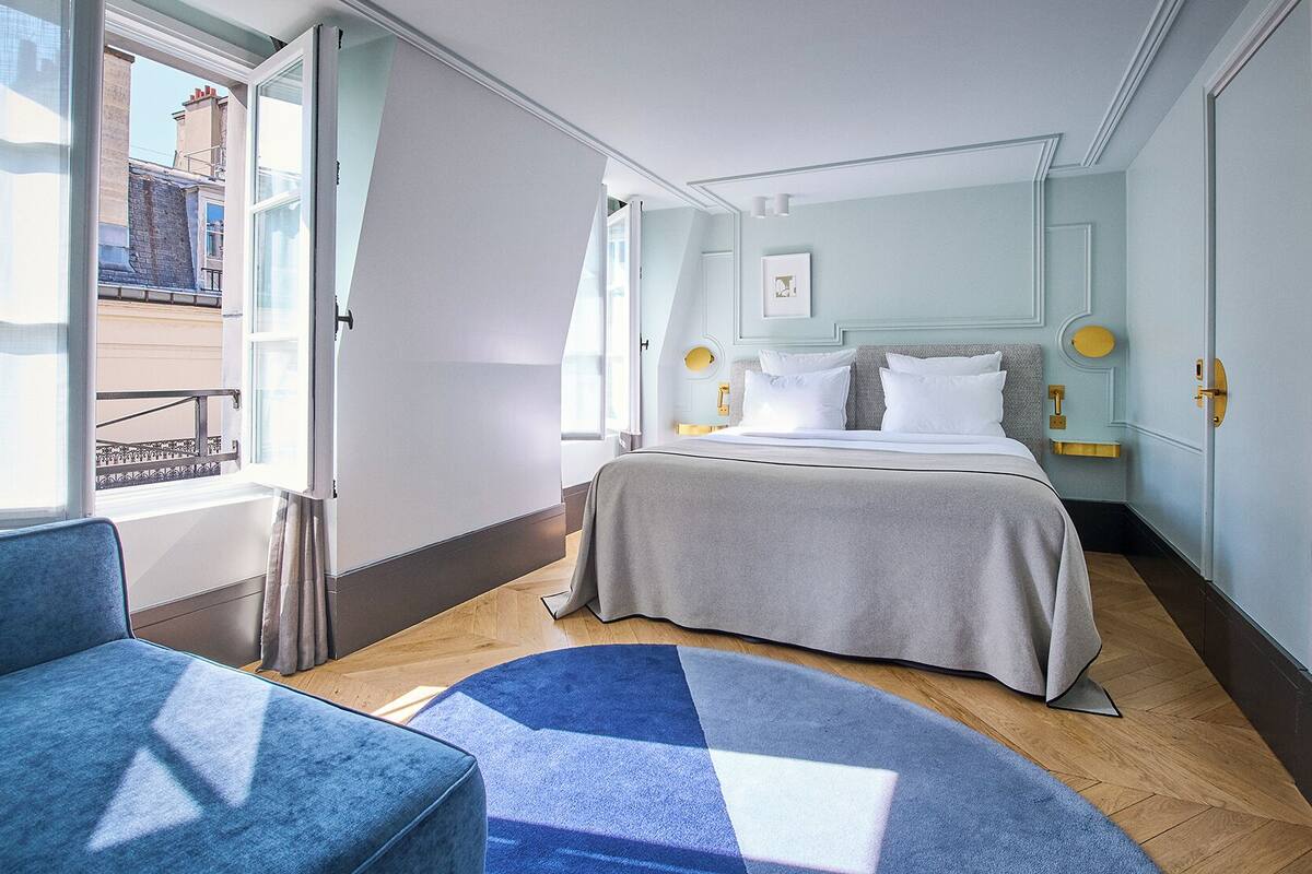 A hotel room with a bed, a chair, a rug, and a window with a view of buildings outside.