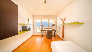 Una habitación compacta con piso de madera, una cama, un escritorio con silla, una mesita con un florero y vistas a montañas a través de ventanas amplias.