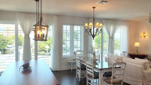 A dining area with a long table, white chairs, and a chandelier. There are windows with curtains, a sofa, and a lamp on a side table.