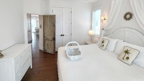 A bedroom with a bed, a nightstand, a white dresser, a ceiling fan, a door, and a window with curtains.