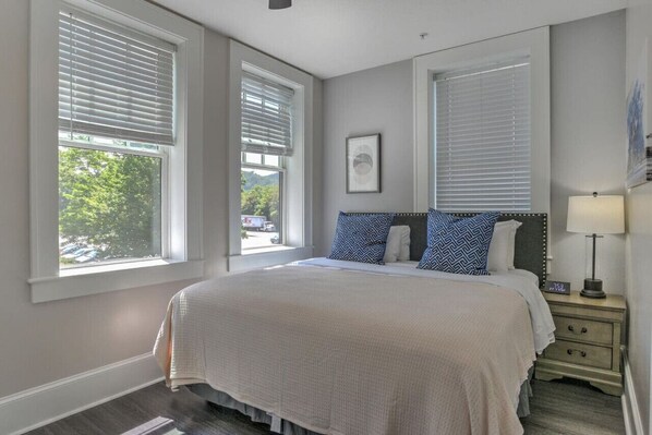 A bedroom with a large bed, two windows with blinds, a bedside table with a lamp, and a framed picture on the wall.