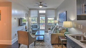 A modern living room with a sofa, chairs, a coffee table, and a TV. There is a ceiling fan, a window with blinds, and a kitchen area with a sink. - Charming Historic King James Building Unit #3 (Asheville)
