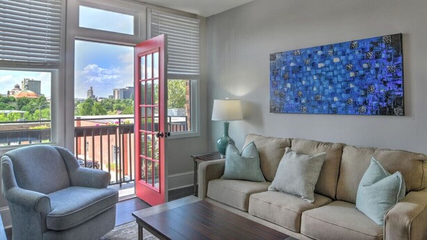 A living room with a sofa, armchair, and a table. There is a window with blinds, a door, and a painting on the wall. - Charming Historic King James Building Unit #3 (Asheville)