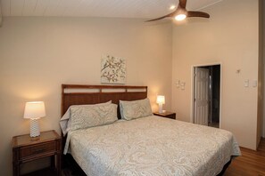 A bedroom with a large bed, wooden headboard, two bedside tables with lamps, a ceiling fan, and a door to another room. - Hale Kamaole 328 Third floor condo, easy access to best beach on Maui! (Kihei)