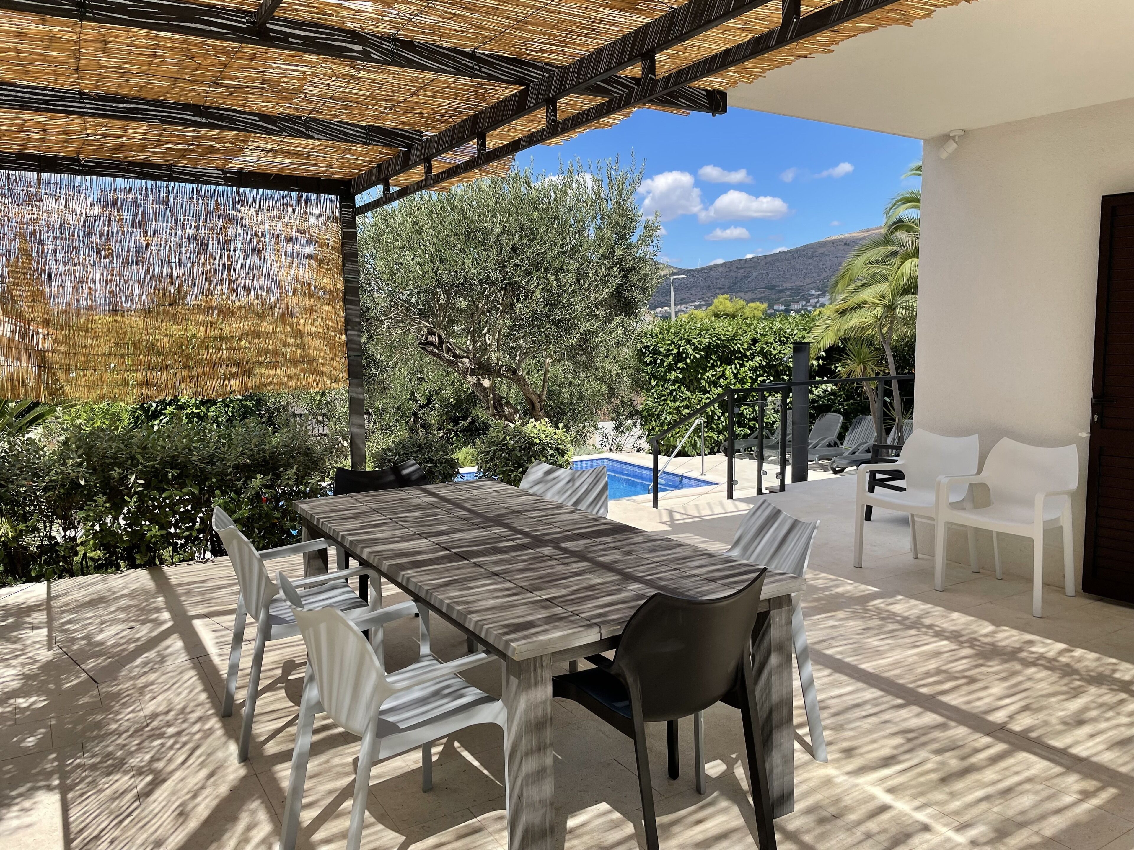 An outdoor patio with a wooden table and chairs, a pool, and a view of the surrounding landscape.