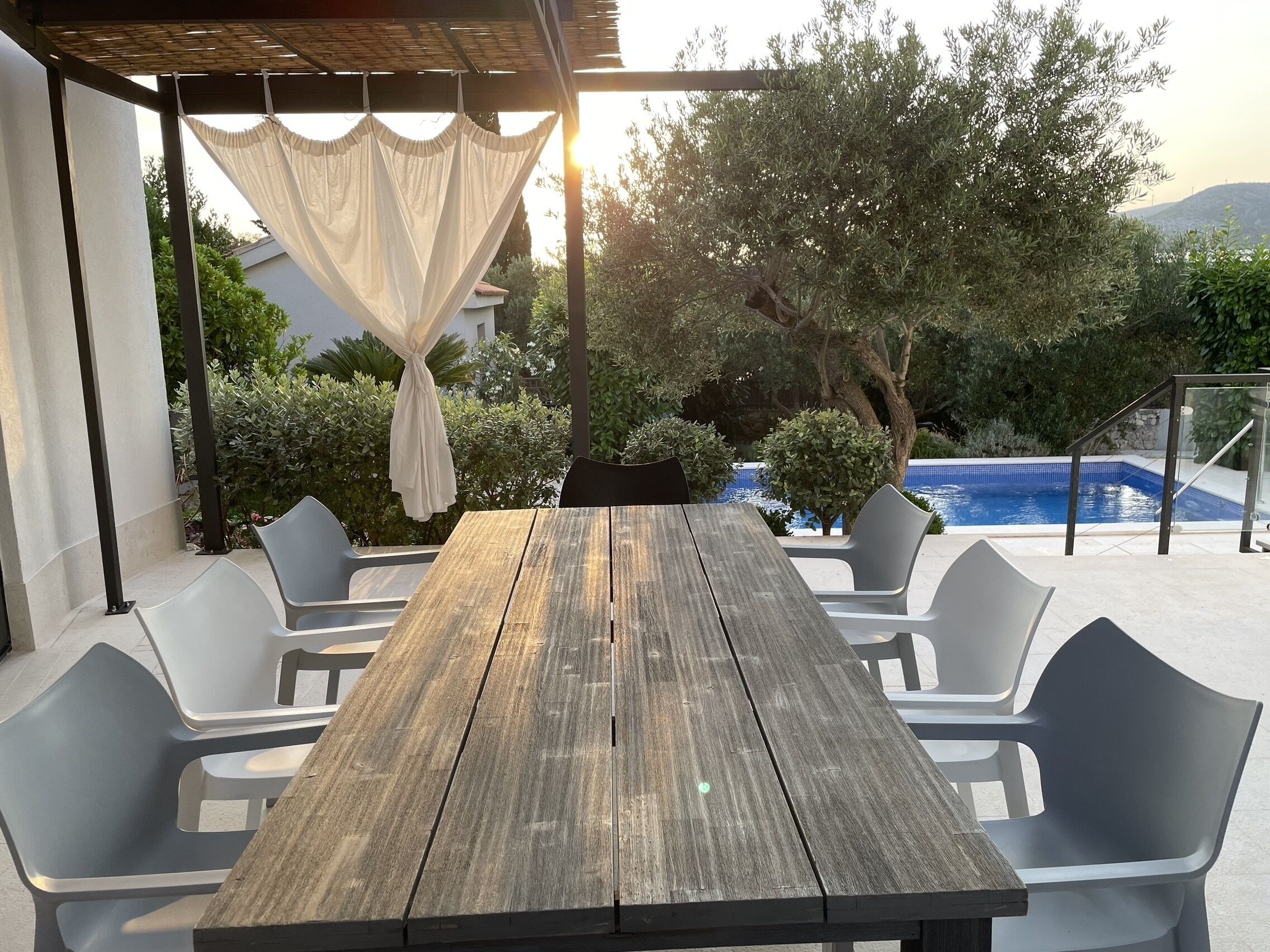 Un tavolo di legno con sei sedie sotto una pergola con tenda bianca, circondato dal verde e da una piscina.