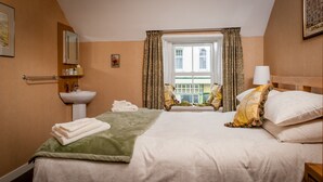 Una habitación de hotel con cama, mesita de noche, lámpara, ventana con cortinas, lavamanos y espejo.