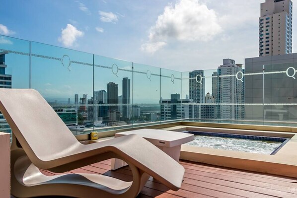 Una terraza en la azotea con una tumbona, un jacuzzi y una pared de vidrio con vista a la ciudad.