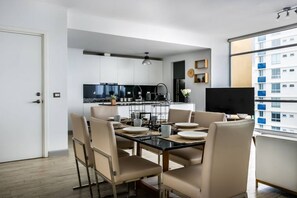 Una cocina moderna con armarios blancos, encimera negra y un comedor con mesa de vidrio y sillas color beige.