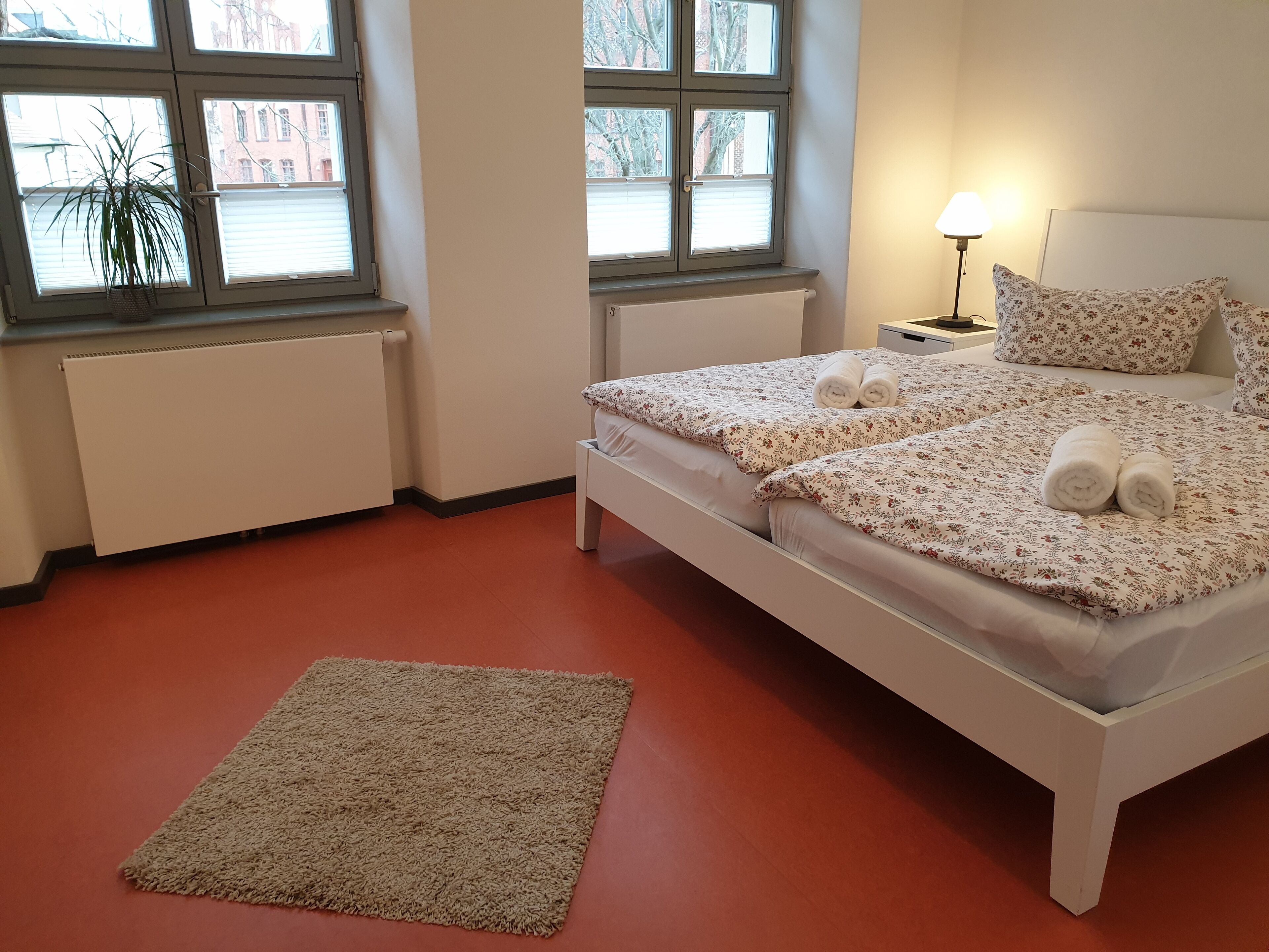 Une chambre à coucher avec un lit, une table de chevet, une lampe, un radiateur et une fenêtre agrémentée d’une plante.