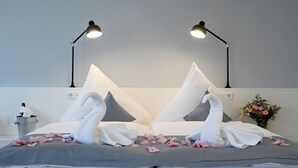 A neatly made bed with white pillows, a grey bedspread, and two swan-shaped towels. There are two bedside lamps, a vase with flowers, and a bucket with a napkin. - Hotel Brandenburger Dom GmbH (Brandenburg an der Havel)