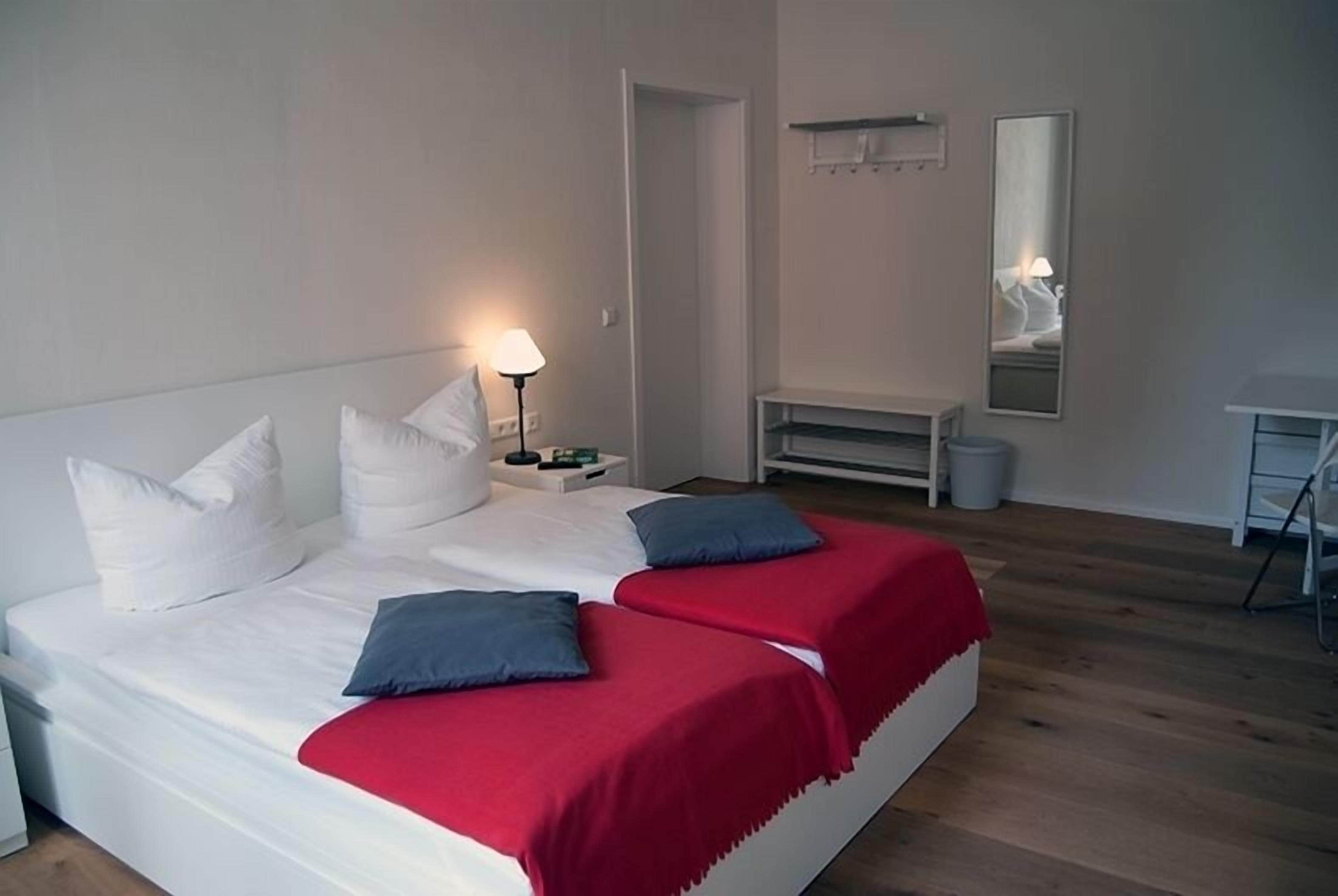 A double bed with white and red bedding, a bedside table with a lamp, a mirror on the wall, and a wooden floor.