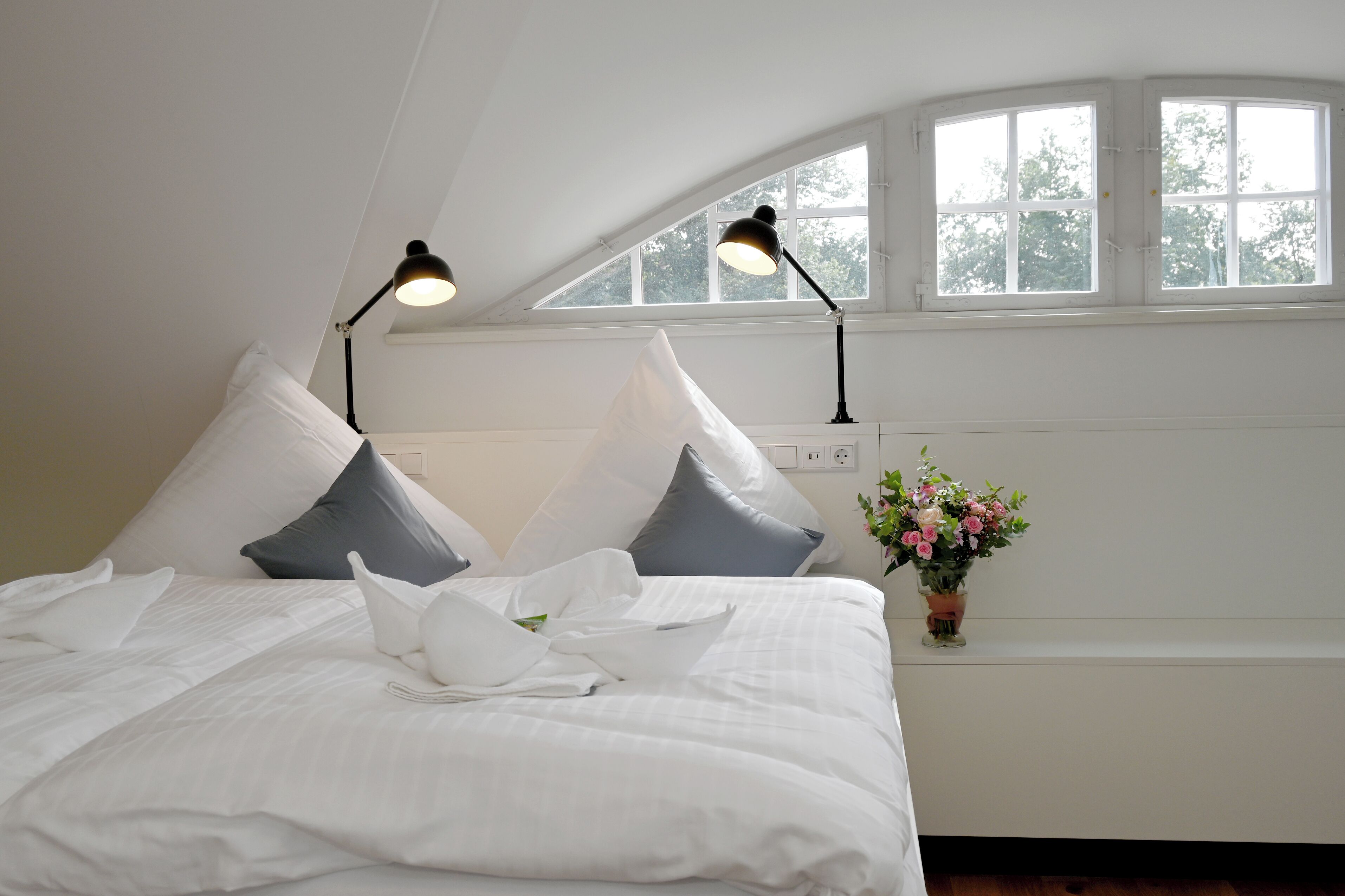 A neatly made bed with white bedding and pillows, two black bedside lamps, a small potted plant with pink flowers, and a window with a view of trees.