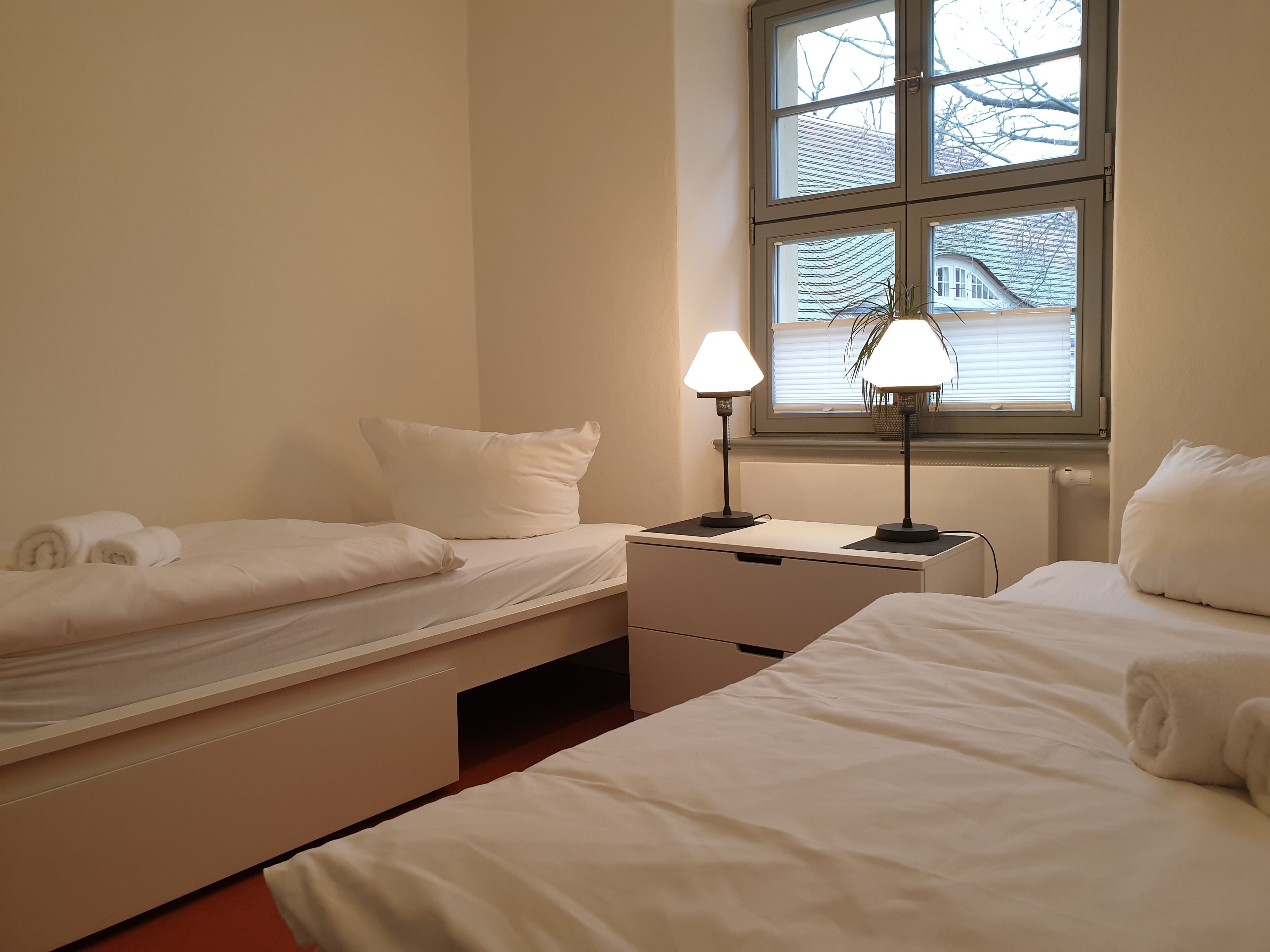 Een hotelkamer met twee bedden, een nachtkastje met een lamp, een raam met jaloezieën en een radiator.