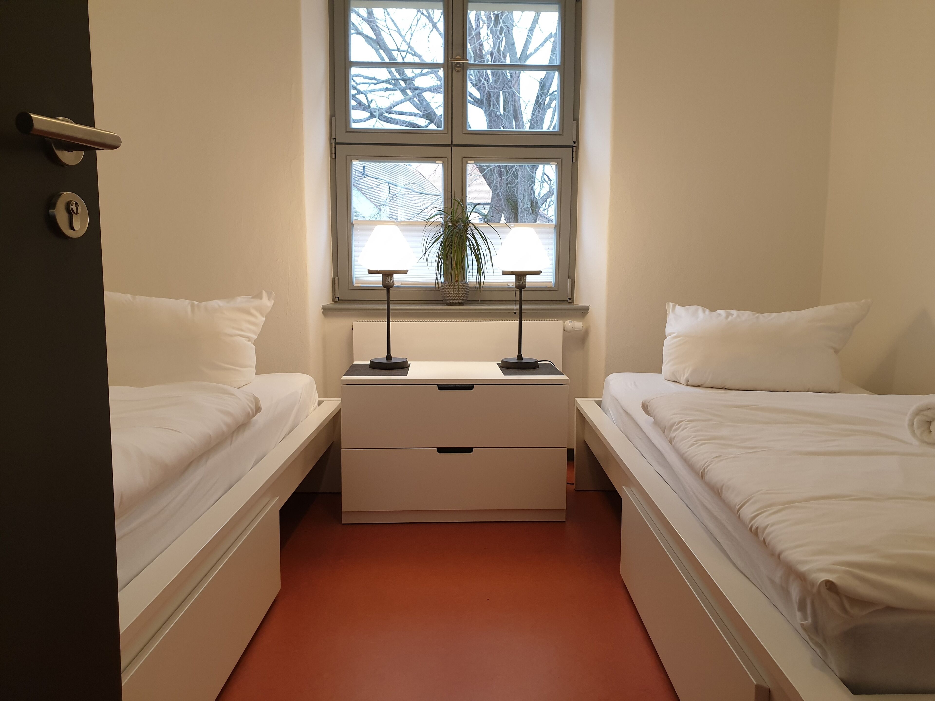 A room with two beds, a nightstand with a lamp, a window with a view of trees, and a door with a lock.