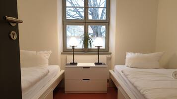 A room with two beds, a nightstand with a lamp, a window with a view of trees, and a door with a lock.